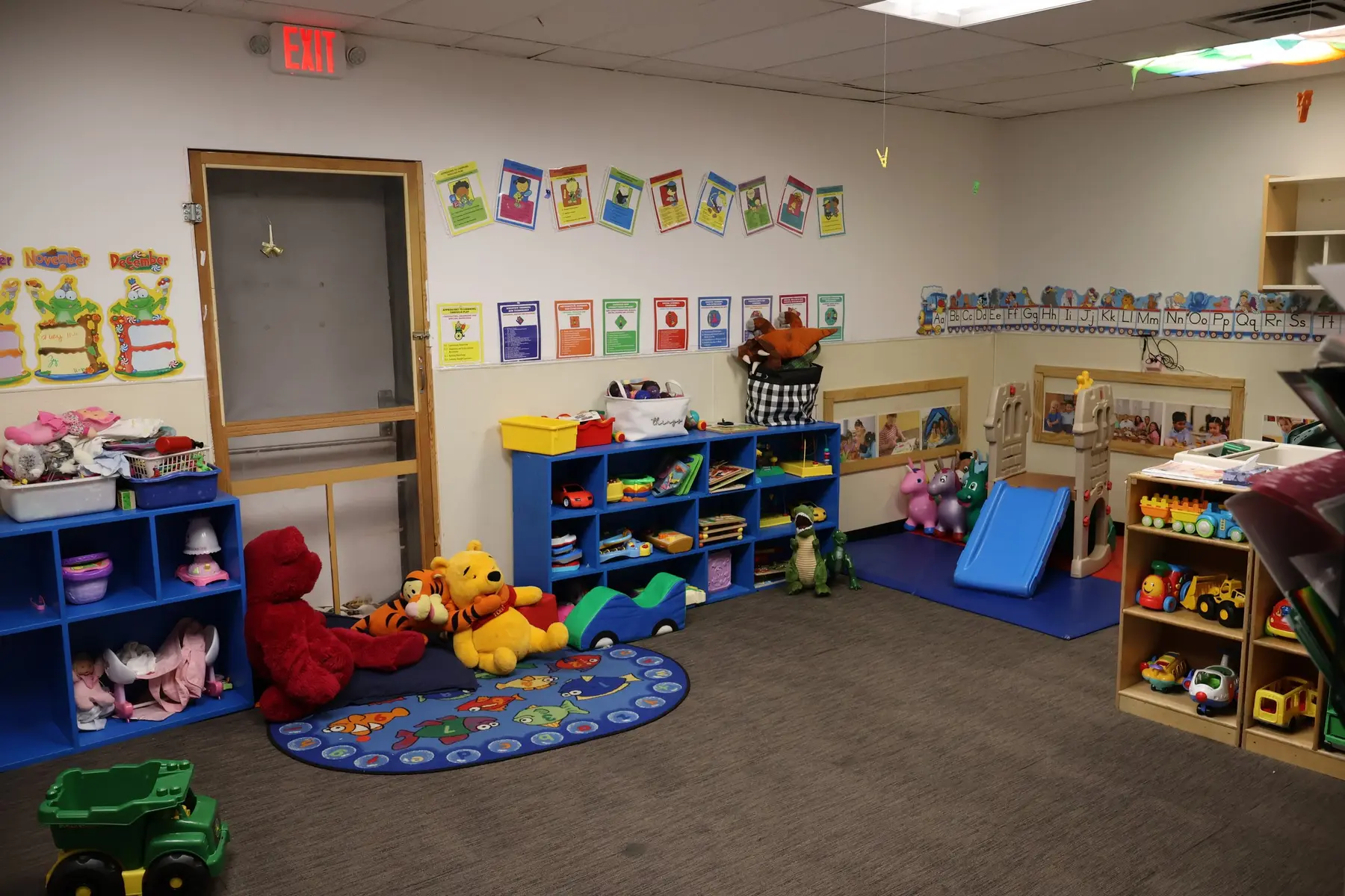 A childcare classroom with blue storage shelves holding toys and stuffed animals, a small blue rug with a road pattern, a play slide, and alphabet posters on the walls.