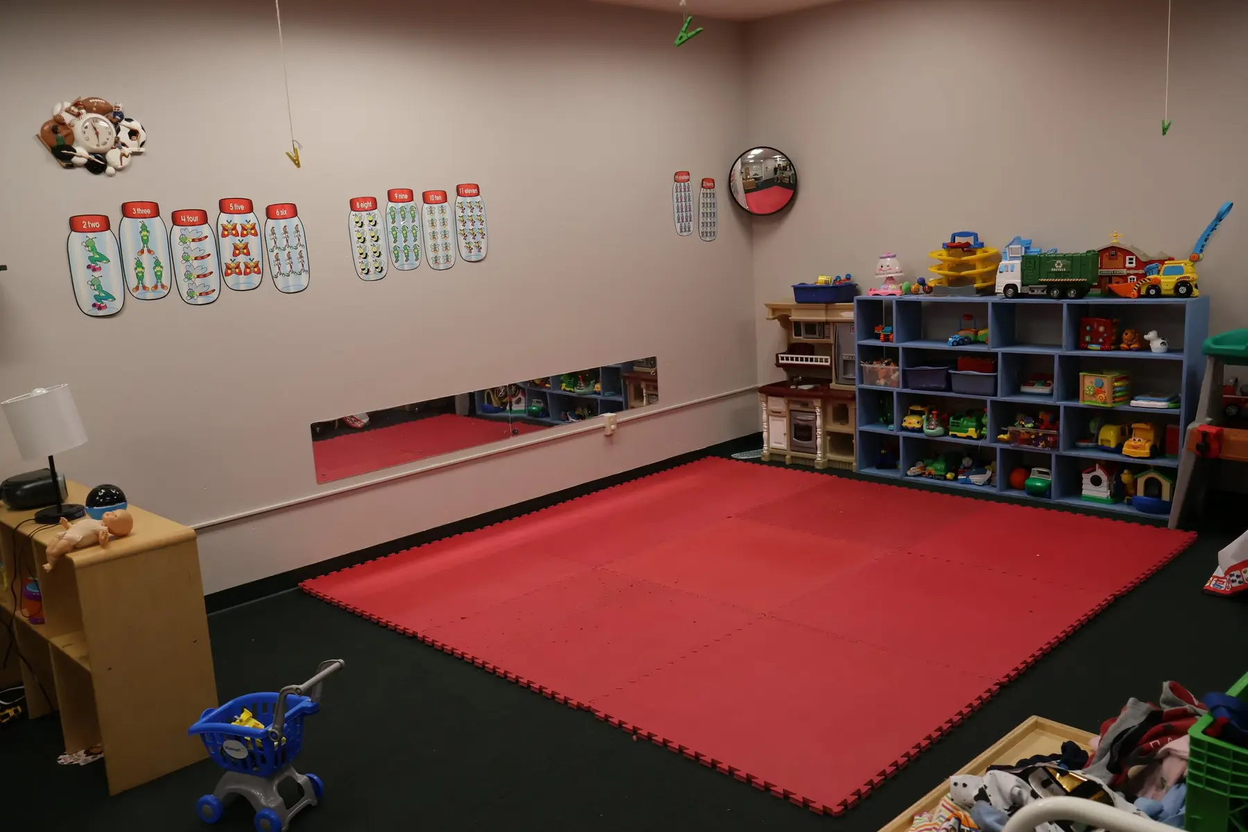 A childcare room with a large red foam mat, blue toy shelves filled with toys, a play kitchen, a small shopping cart, and wall decorations including number posters and a mirror.