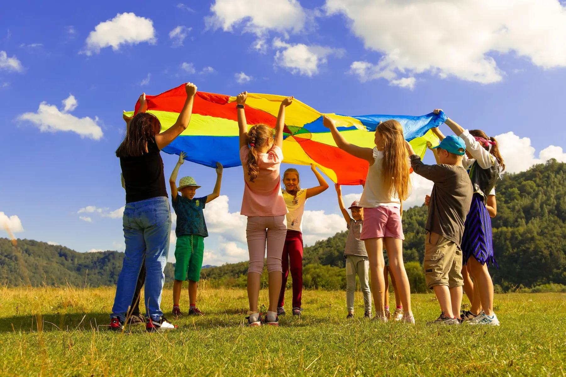 Nine children stand in a grassy field holding the edges of a large multicolored parachute up against a blue sky with clouds.