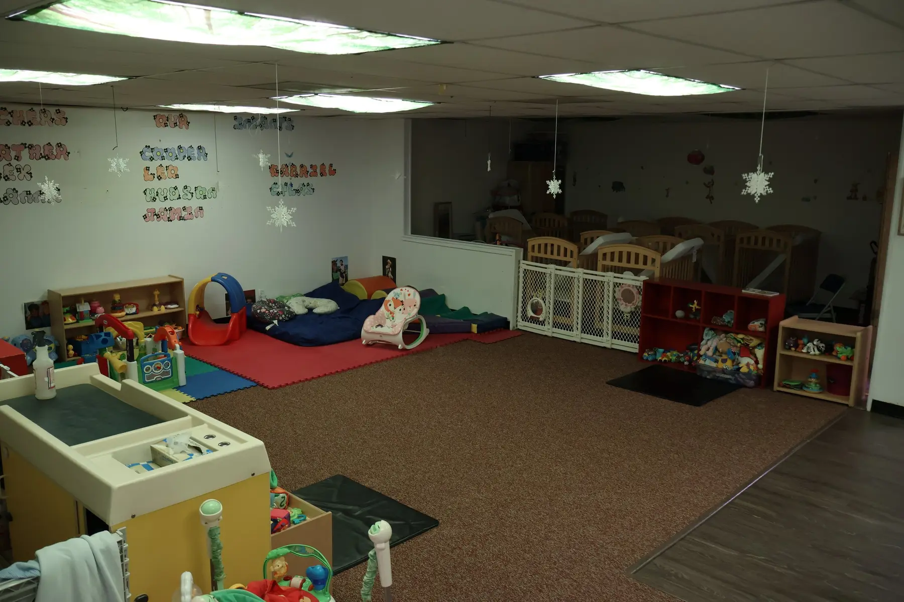 An empty childcare room with a brown carpet, colorful foam mats, a play gym, a changing table, low shelves with toys, and a gated area containing cribs.