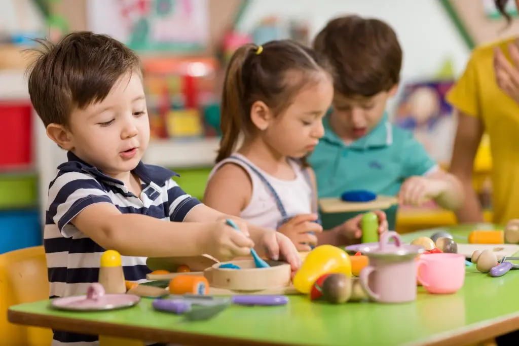 Three young children sit at a green table with wooden toys and colorful objects, engaged in play.