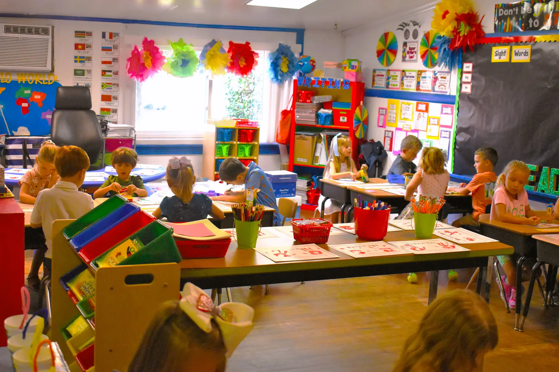 Children seated at desks in a colorful classroom with educational posters and craft supplies; some are writing or drawing on paper.