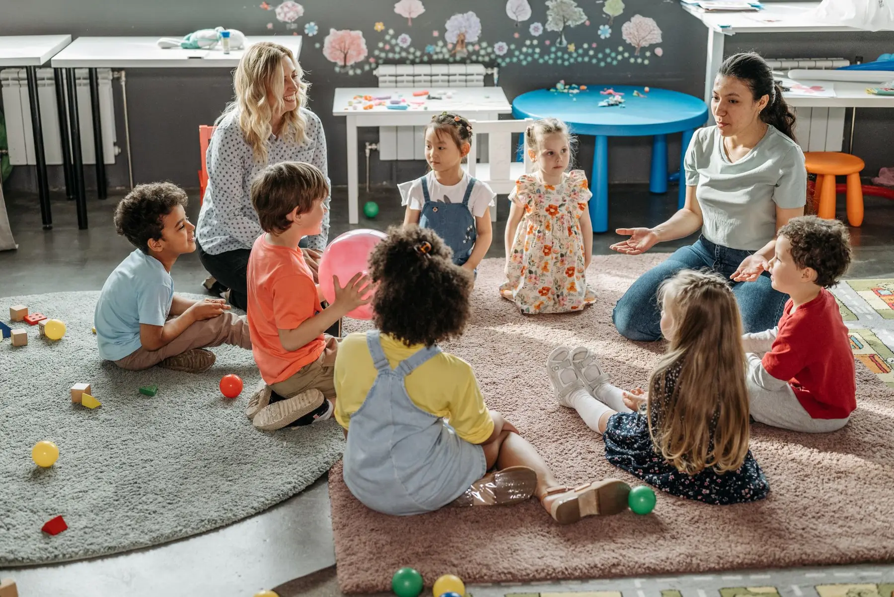 Eight children and two adults sit in a circle on rugs with toys and balls around them in a classroom setting.