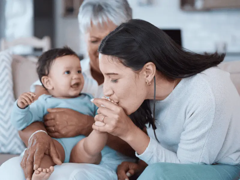 A woman kisses the hand of a baby held by an older woman on a couch.