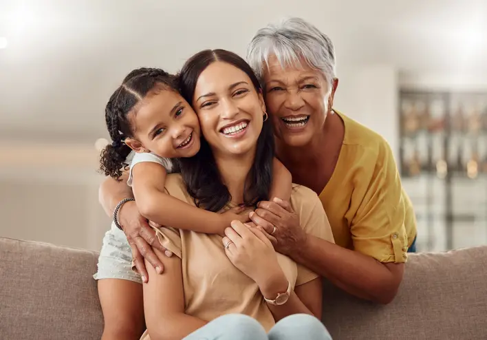 Three smiling people sit together on a sofa, with a child hugging a woman from the side and an older woman embracing her from behind.