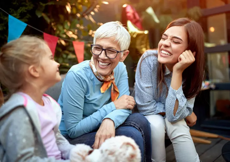 Two women and a young girl sit together outdoors, smiling and laughing, with a small fluffy dog in the foreground and colorful bunting overhead.