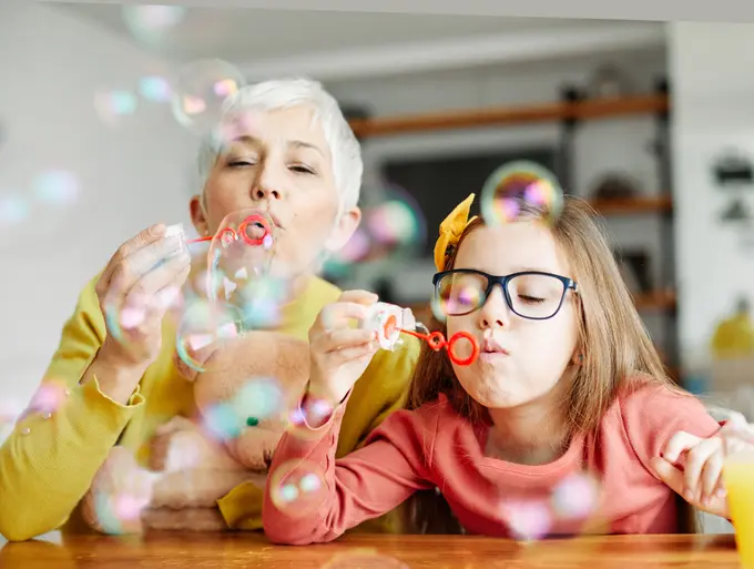 An older woman and a young girl blow bubbles through wands at a wooden table, with a teddy bear resting near the woman's arm.