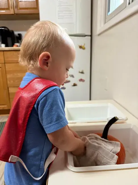 A young child wearing a red cape and blue shirt stands at a kitchen sink washing a pumpkin with a cloth.