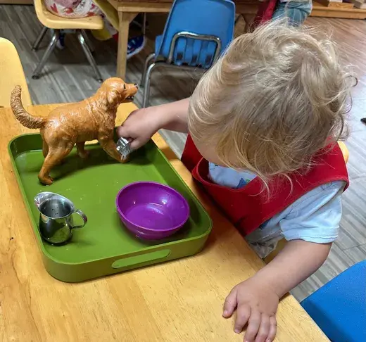 A young child with blond hair leans over a green tray, holding a small metal cup near a brown toy dog, with a purple bowl resting on the tray.