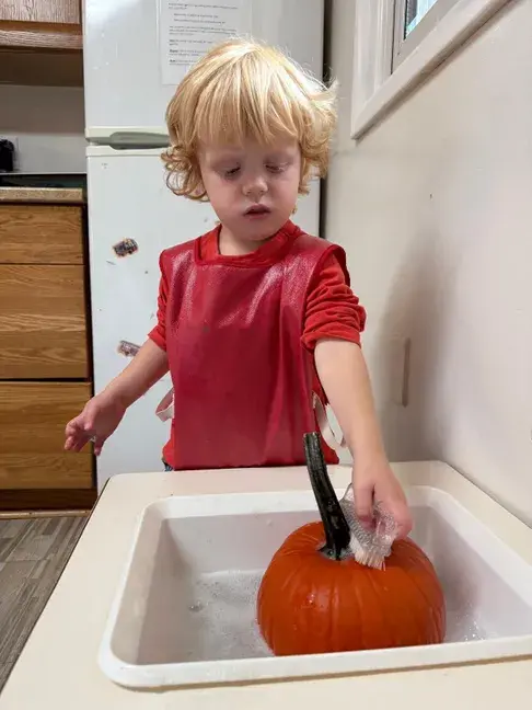 A young child in a red shirt stands at a kitchen sink washing a small orange pumpkin with a scrub brush.