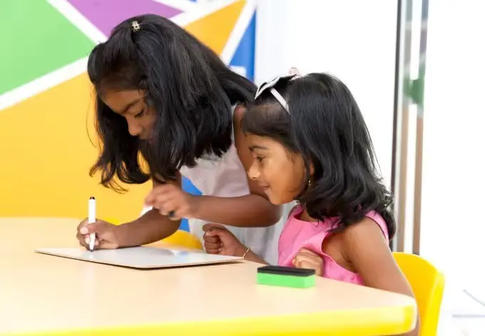Two girls sit at a yellow table, one writing on a whiteboard with a marker while the other watches, with a green eraser nearby.