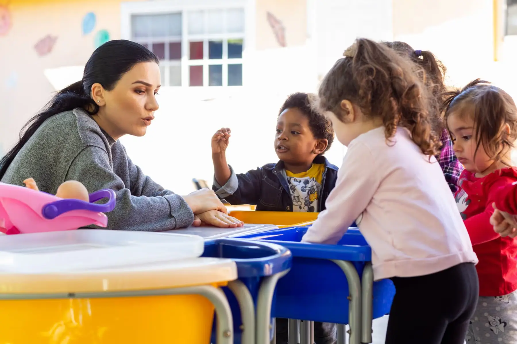 A woman leans over a table talking with a young boy while two other children stand nearby in a play area.