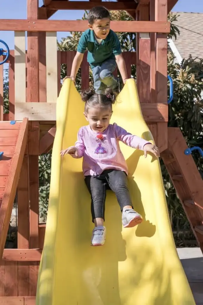 A girl in a pink shirt slides down a yellow slide while a boy in a green shirt stands on the wooden play structure behind her.