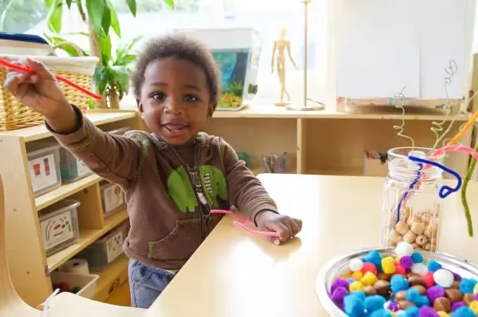 A young child sits at a table holding a red pipe cleaner in one raised hand and a pink pipe cleaner in the other, with a tray of colorful pom-poms and a jar of beads nearby.