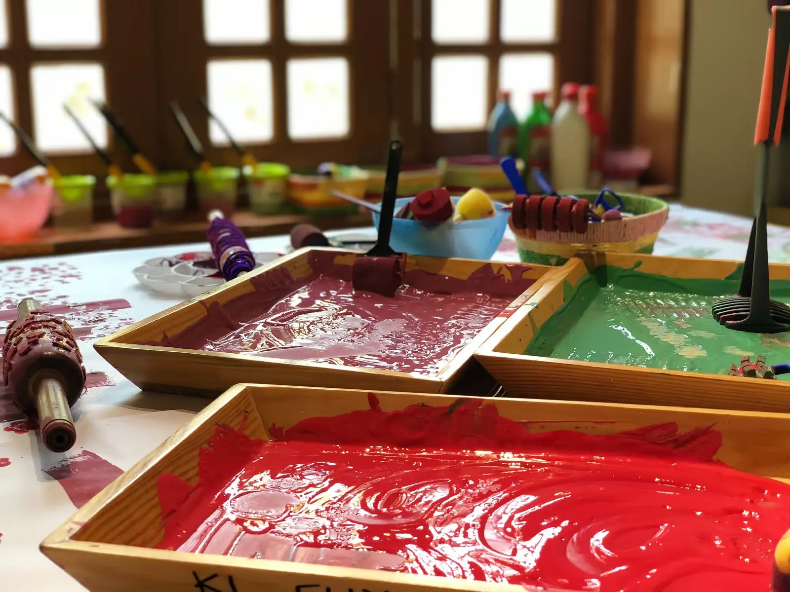 Three wooden trays filled with red, purple, and green paint sit on a paint-splattered table alongside paint rollers and brushes.