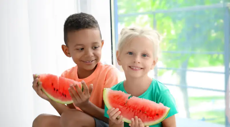 Two children sit side by side holding large slices of watermelon and smiling.