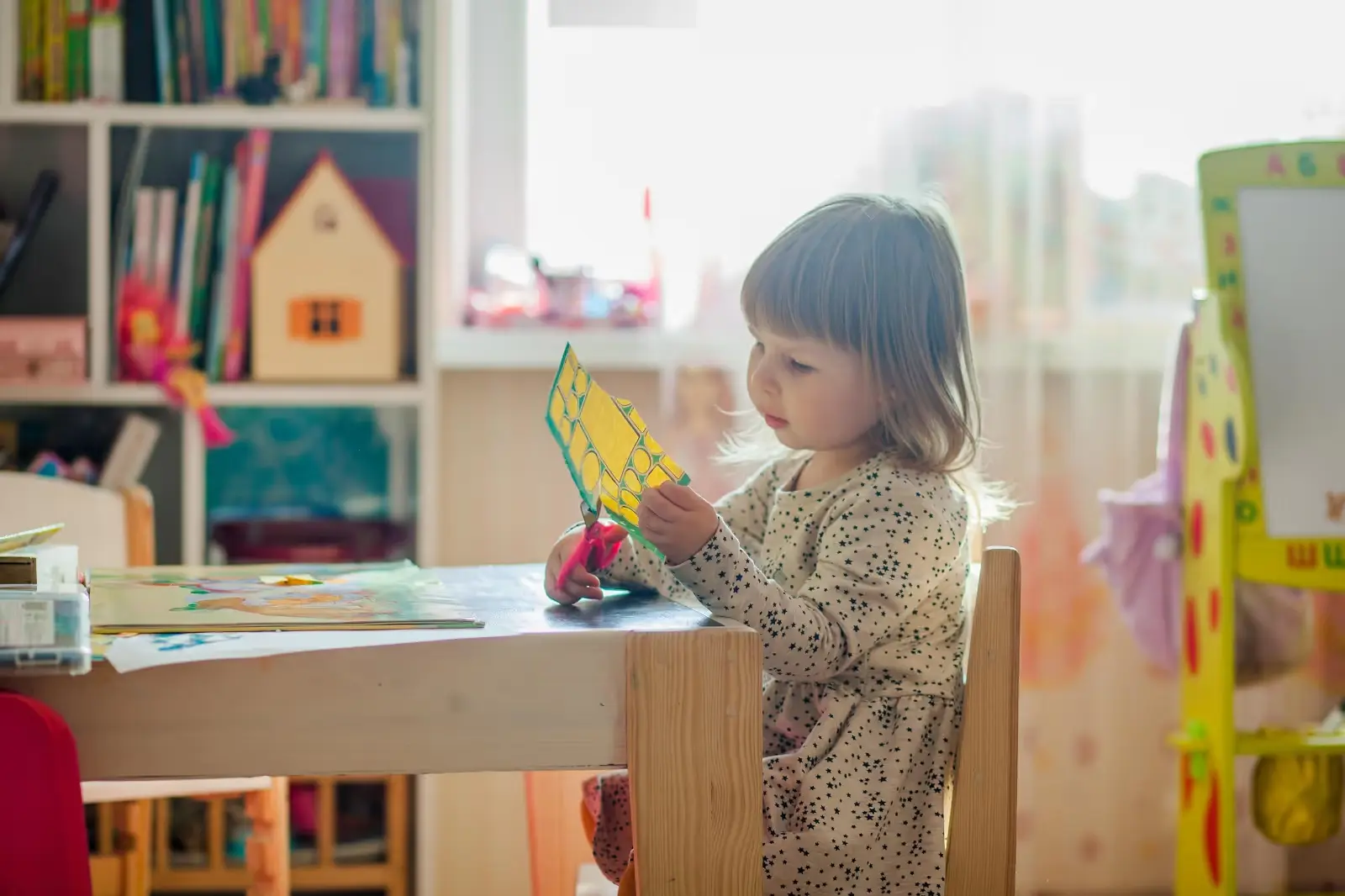 A young girl in a polka-dot dress sits at a table holding a yellow paper craft with green shapes.