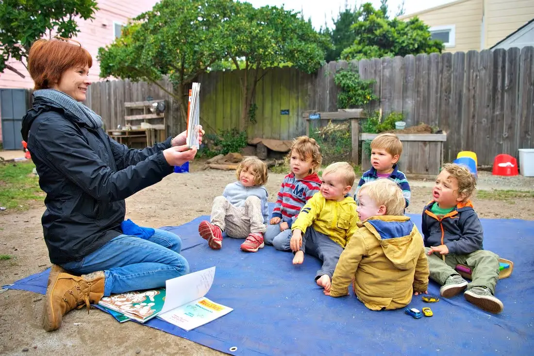 A woman sits on a blue mat holding a book while six young children sit facing her in an outdoor play area with a wooden fence and play structures.