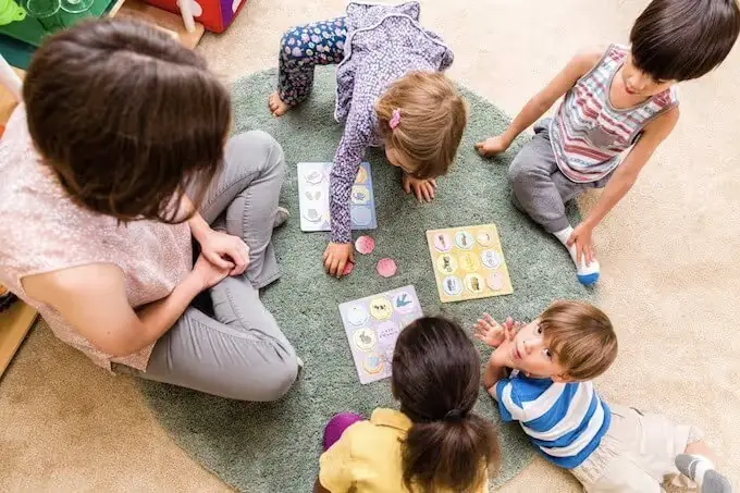 A woman sits on a green rug with four children who are playing with circular game pieces and printed boards.