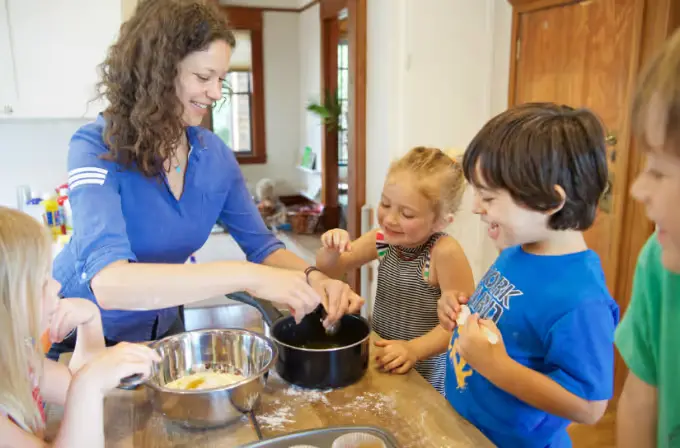 A woman in a blue shirt helps four children mix ingredients in bowls on a kitchen counter.