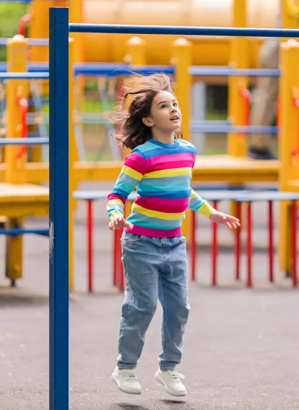 A child in a colorful striped shirt and jeans jumps near playground equipment.