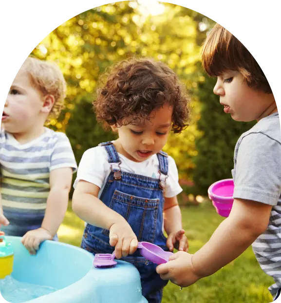 Three toddlers play with water in a blue plastic tub outdoors using purple and pink scoops.