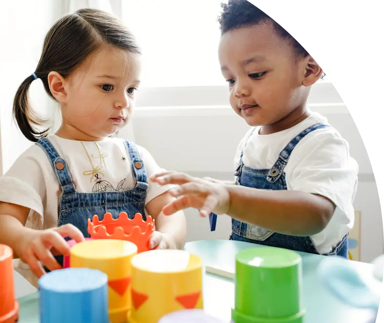 Two toddlers in overalls interact with colorful stacking cups on a table.
