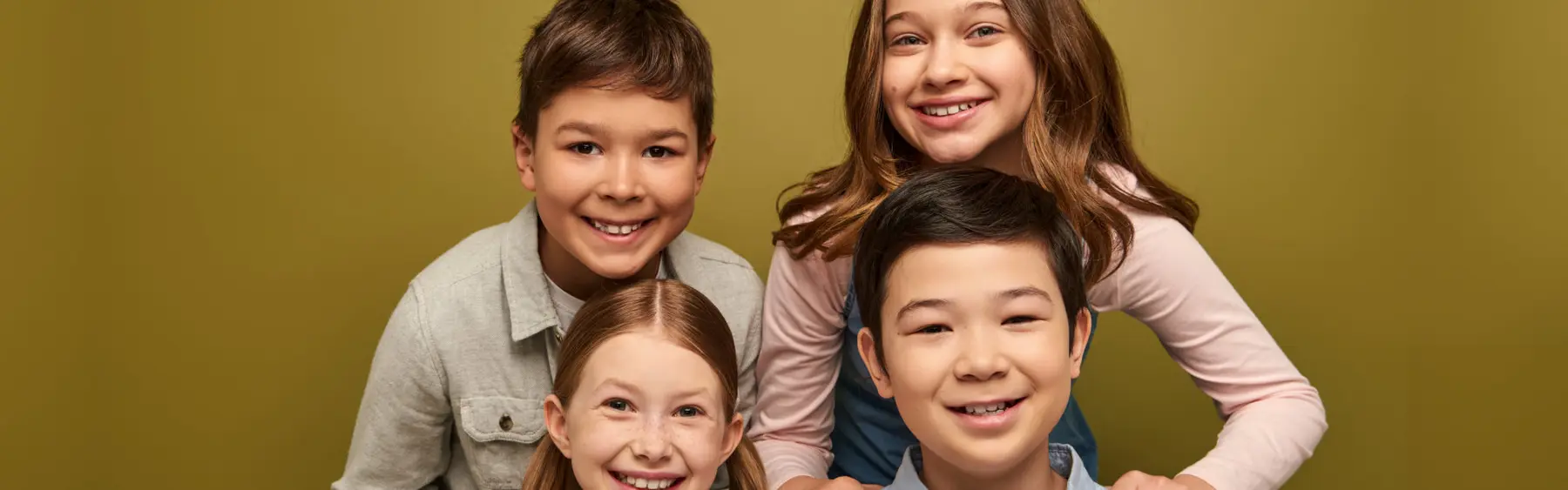 Four smiling children posing closely together against a solid olive-green background.