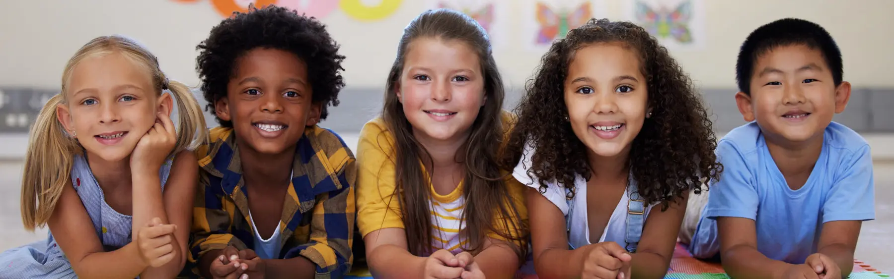 Five diverse children sitting together on a colorful mat in a classroom setting with butterfly decorations on the wall.