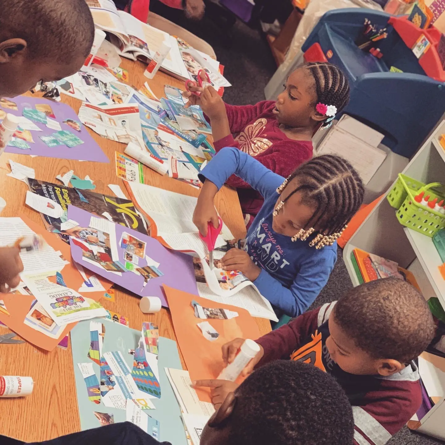 Children sit at a table cutting and gluing magazine clippings onto colored paper.