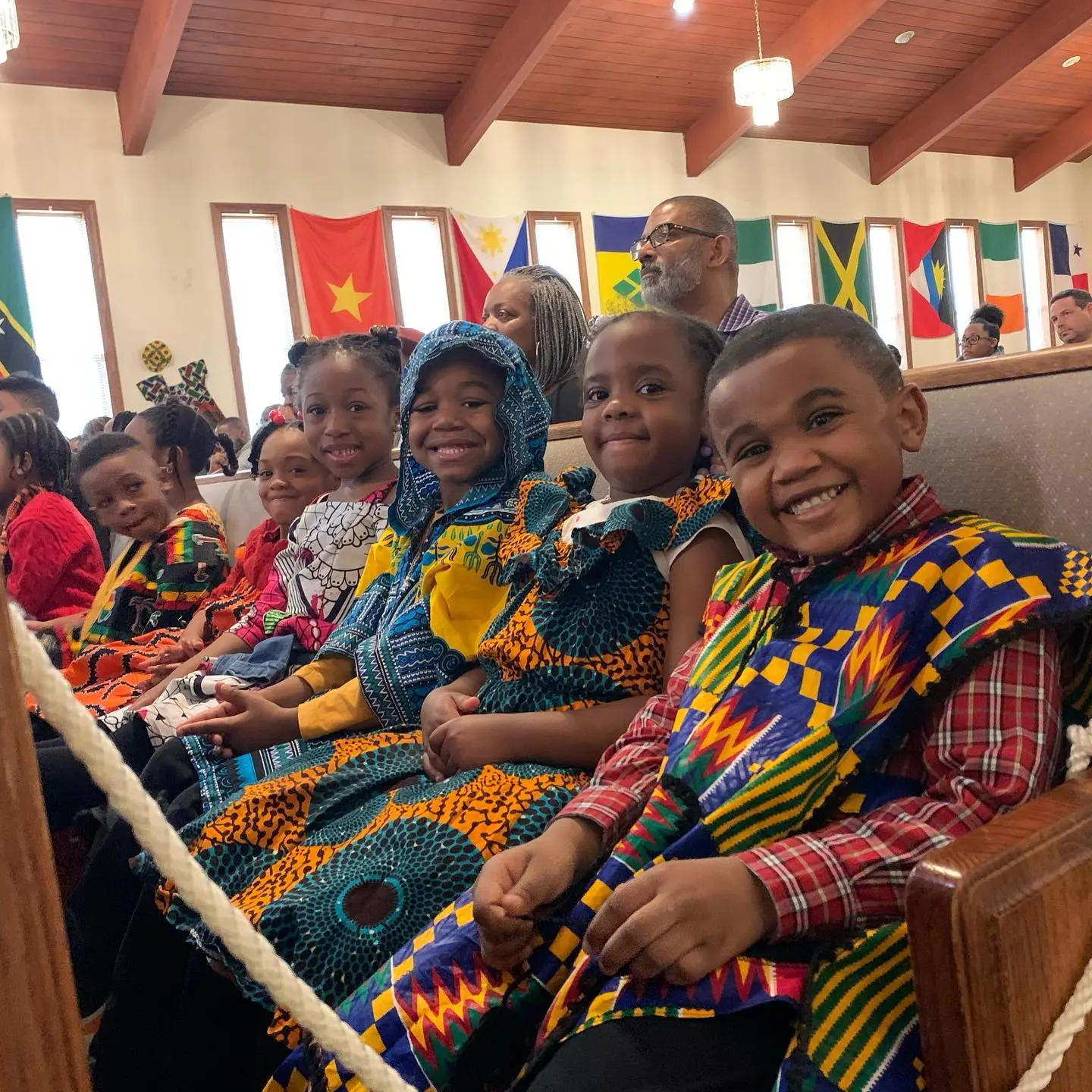 Children wearing colorful patterned clothing sit in wooden pews while adults sit behind them in a room decorated with multiple national flags.