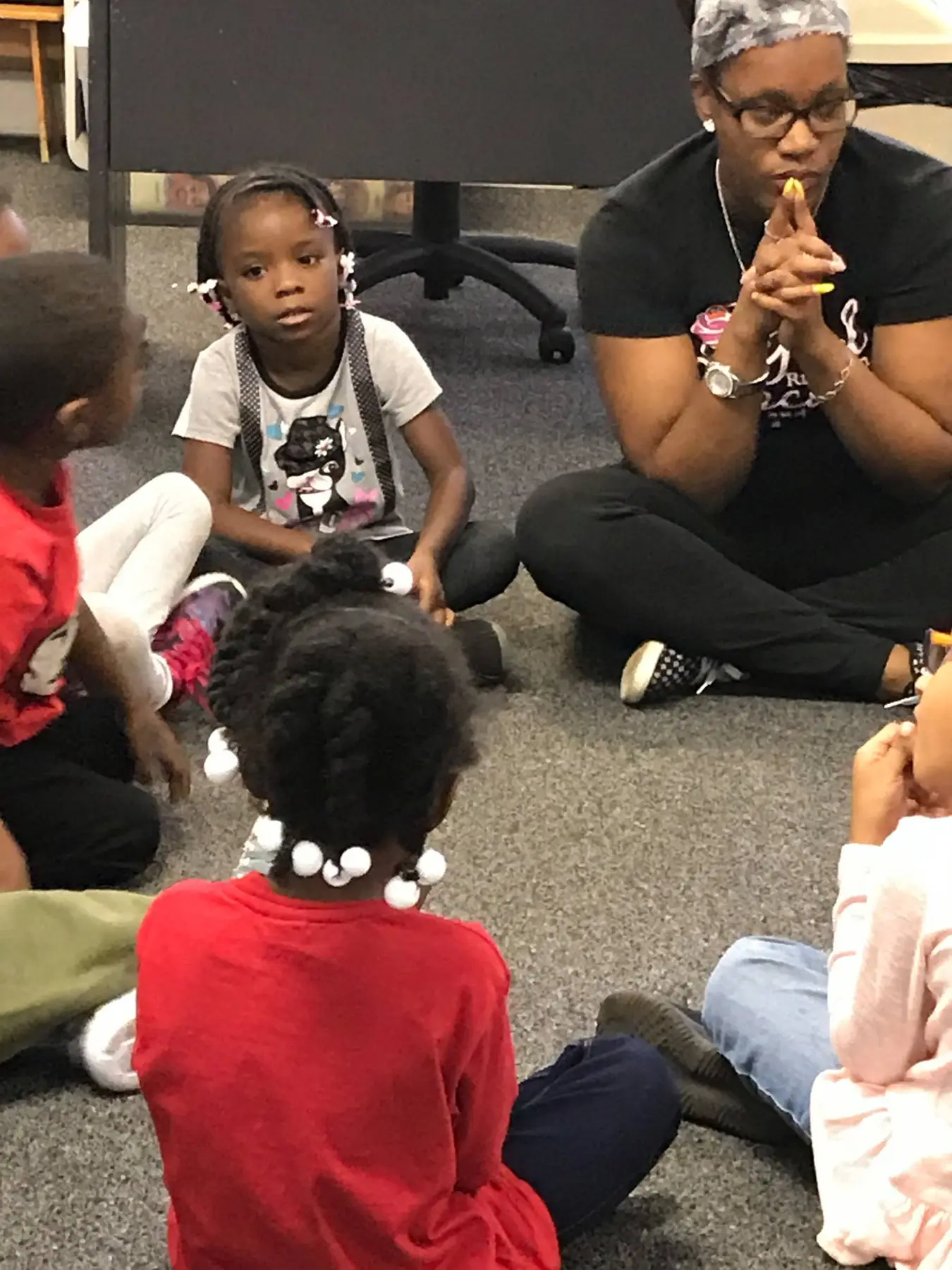 An adult woman sits cross-legged on a carpeted floor with hands clasped, facing four children seated in a circle around her.