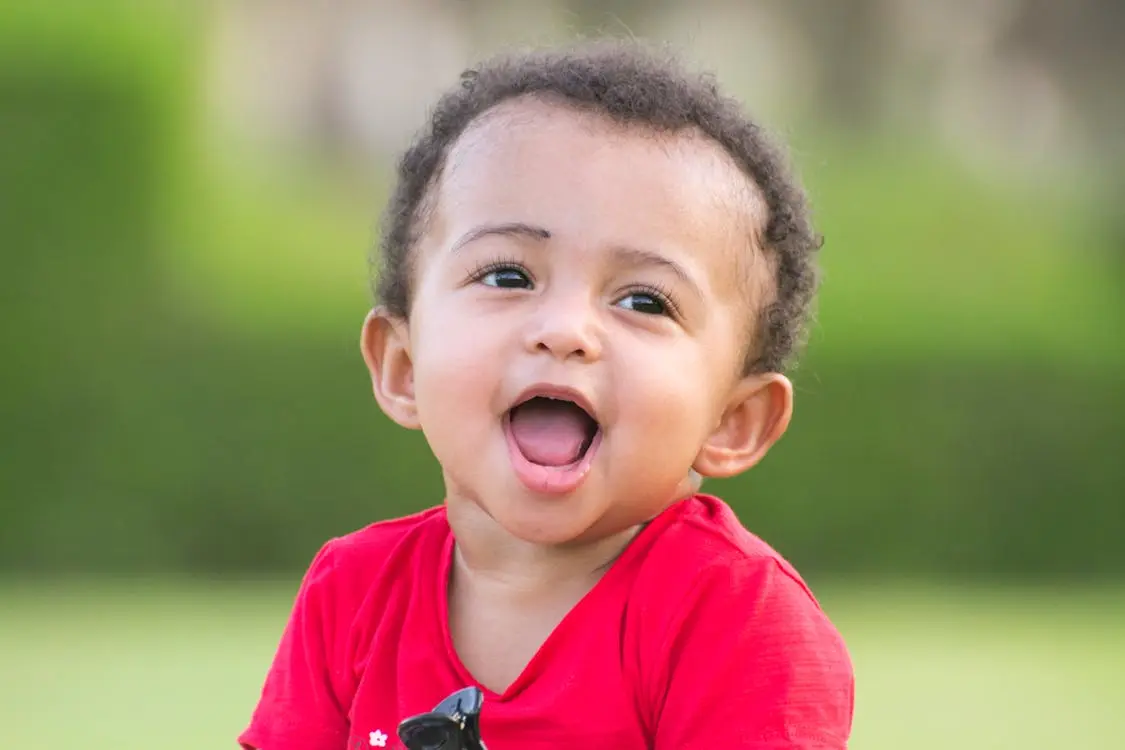 A smiling toddler in a red shirt with sunglasses hanging from the neckline looks upward against a blurred green outdoor background.