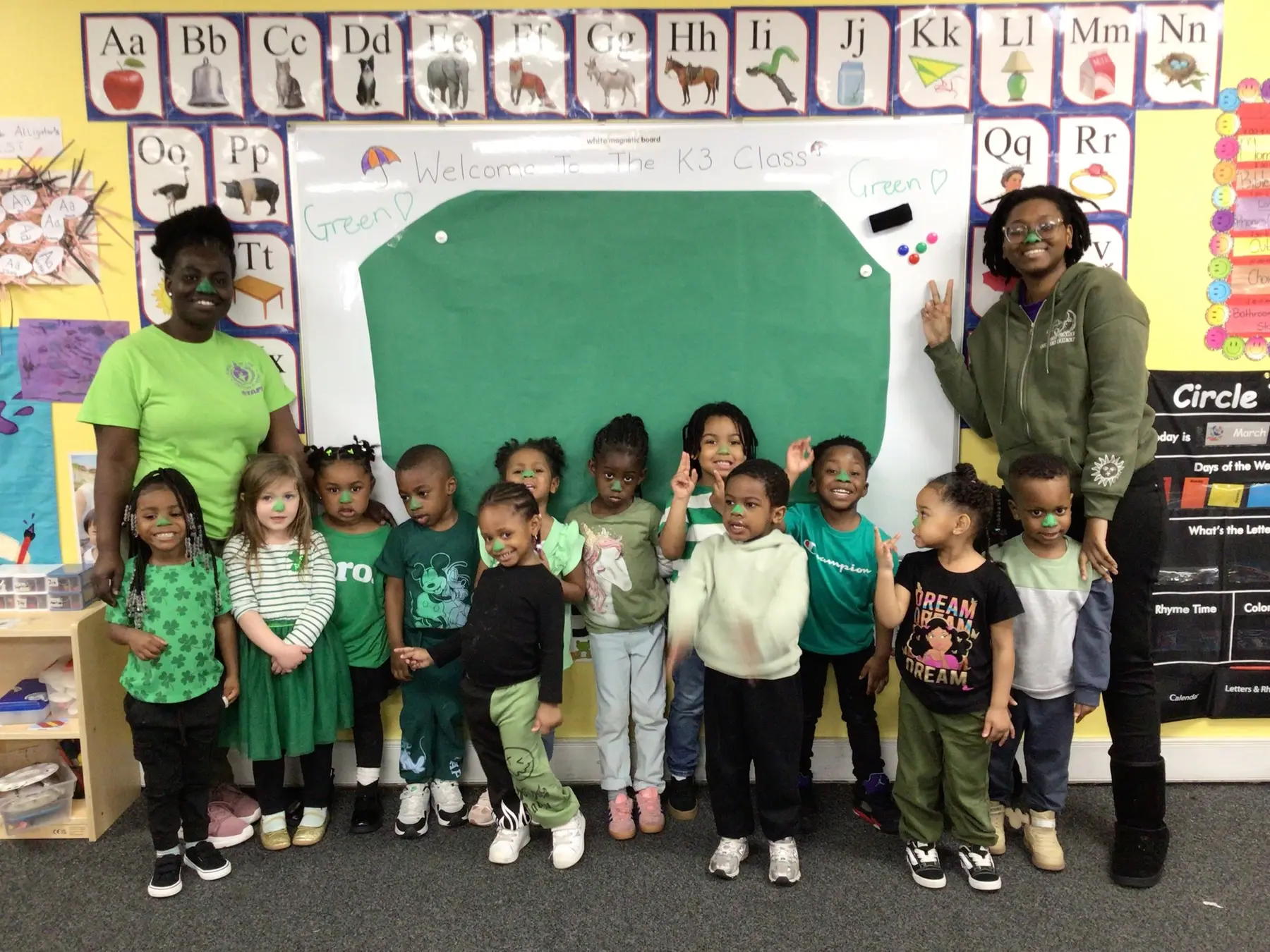 Two adults stand beside a group of eleven children, most wearing green, in front of a whiteboard and alphabet posters in a classroom.