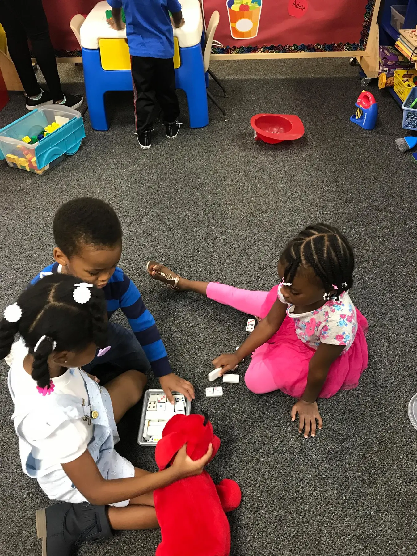Three children sit on a carpeted floor playing with blocks and a red stuffed animal while another child stands at a blue table in the background.