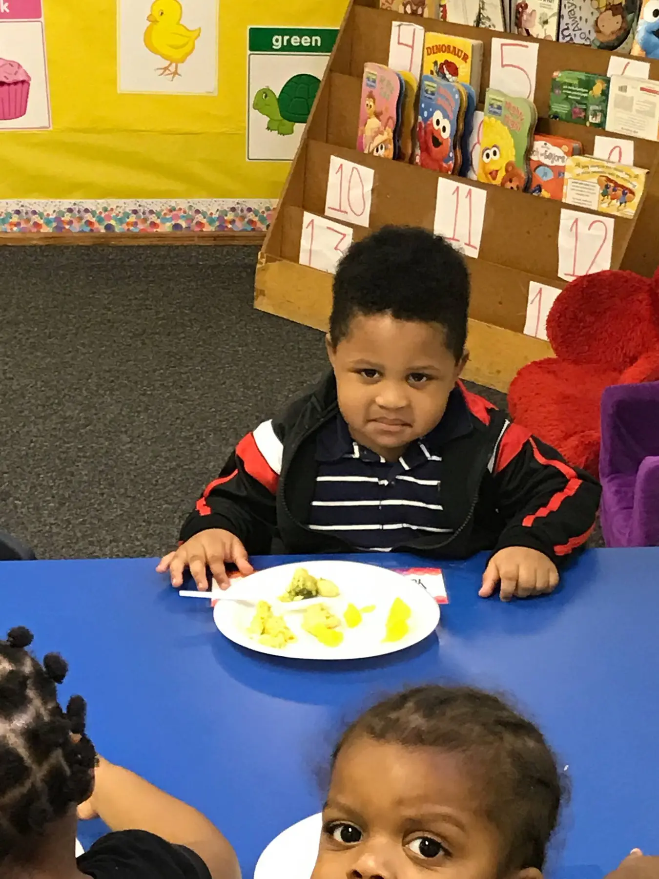 A boy in a striped shirt and black jacket sits at a blue table with a plate of food while two other children are partially visible in the foreground.