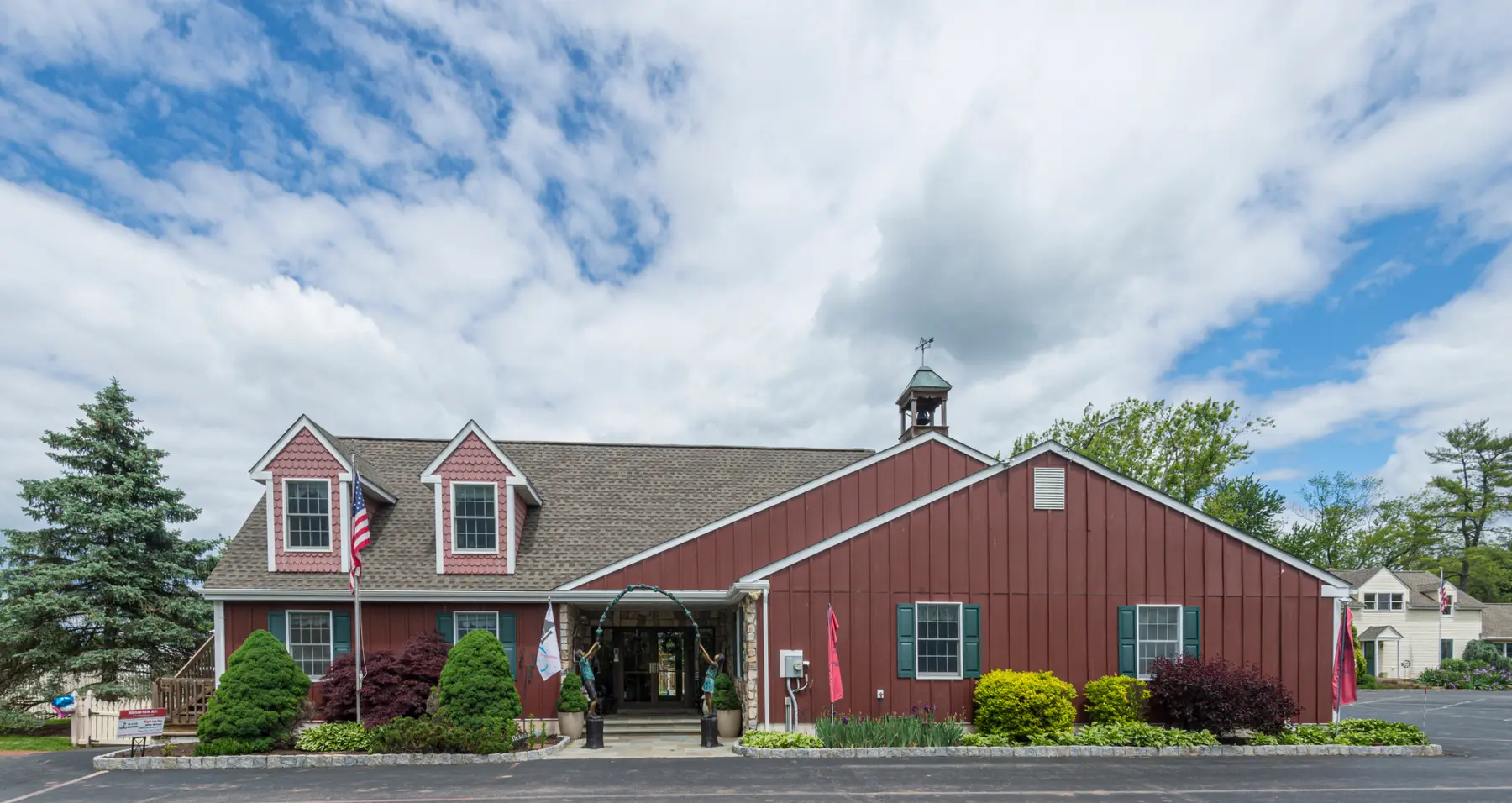 A red wooden building with a gray shingled roof, dormer windows, and a small cupola stands behind a paved parking area and landscaped bushes under a cloudy blue sky.