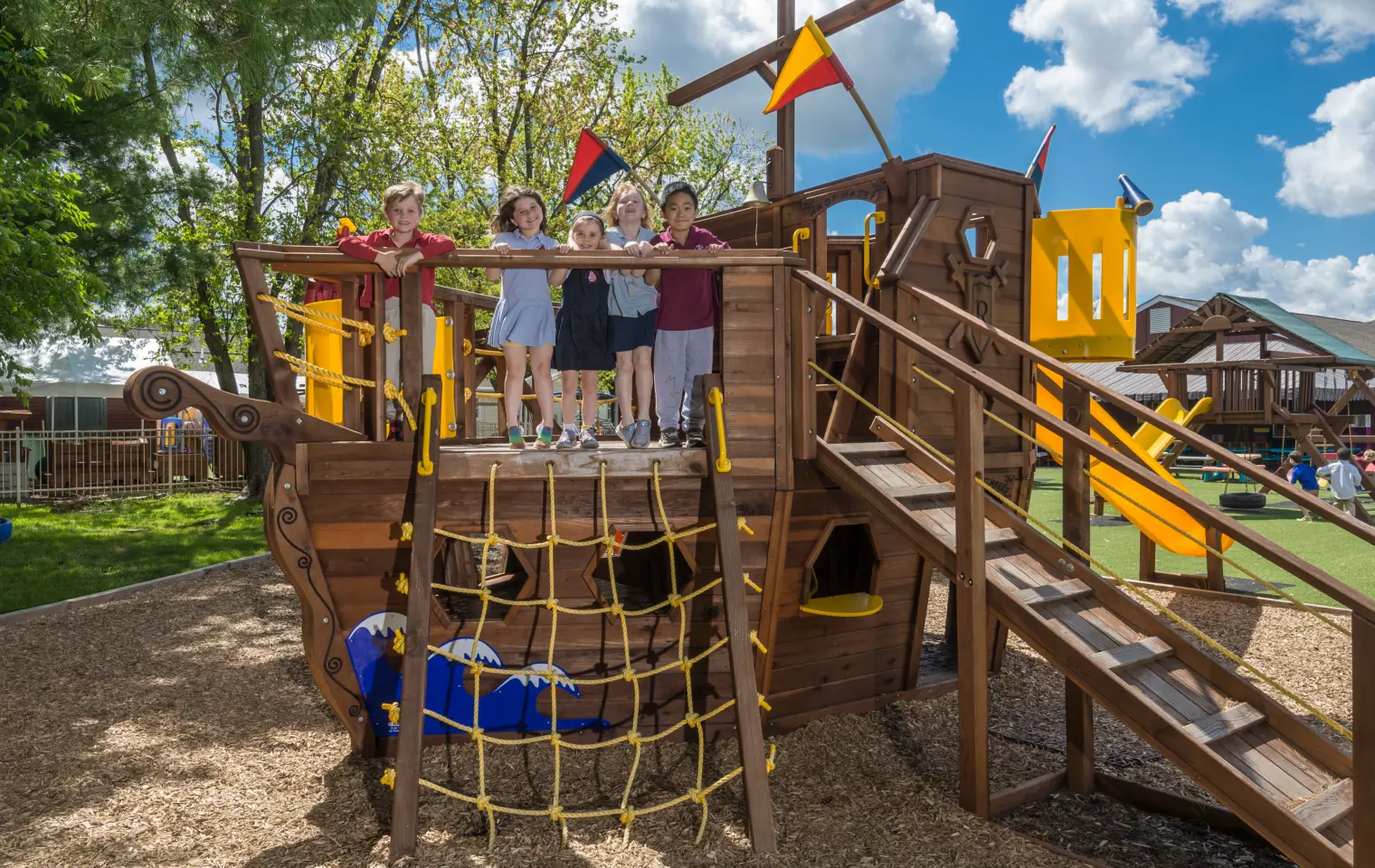 Five children stand on the deck of a wooden pirate ship play structure with a yellow rope net, wooden stairs, and flags, set on a mulch-surfaced playground under a blue sky.