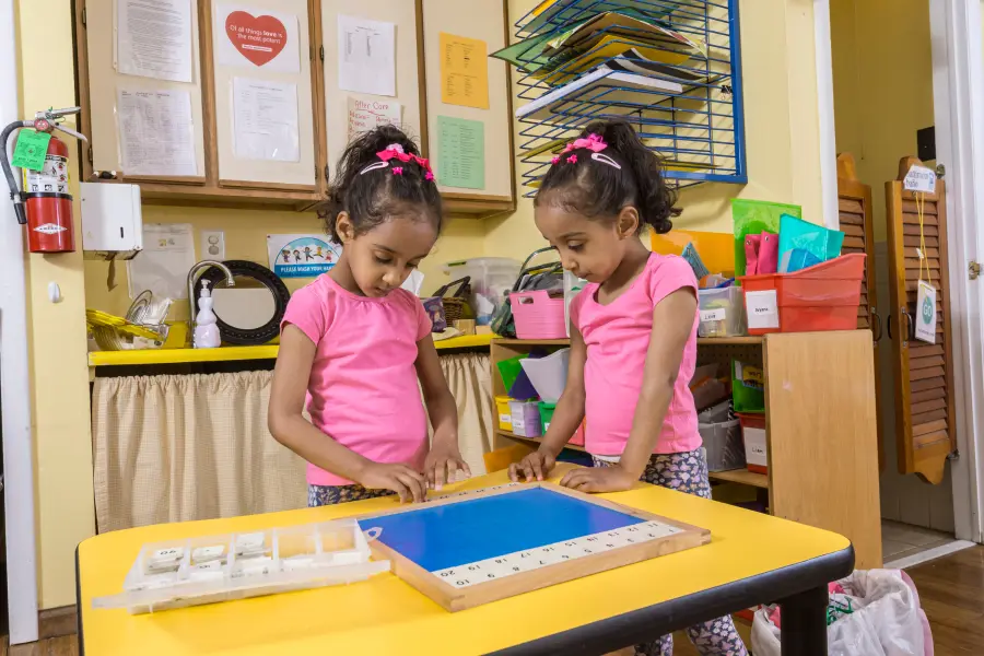 Two girls in pink shirts stand at a yellow table, one touching a blue tray with numbered tiles while the other watches, in a room with cabinets, shelves and a fire extinguisher.