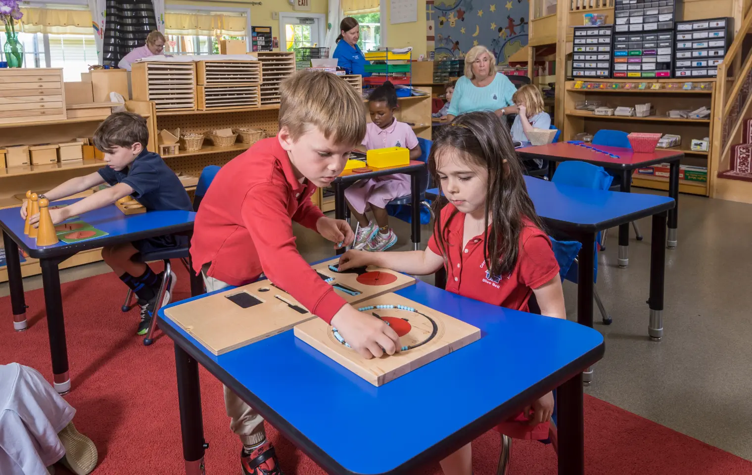 Children work with wooden shape puzzles at blue tables while adults stand and sit nearby in a classroom with shelves of materials.