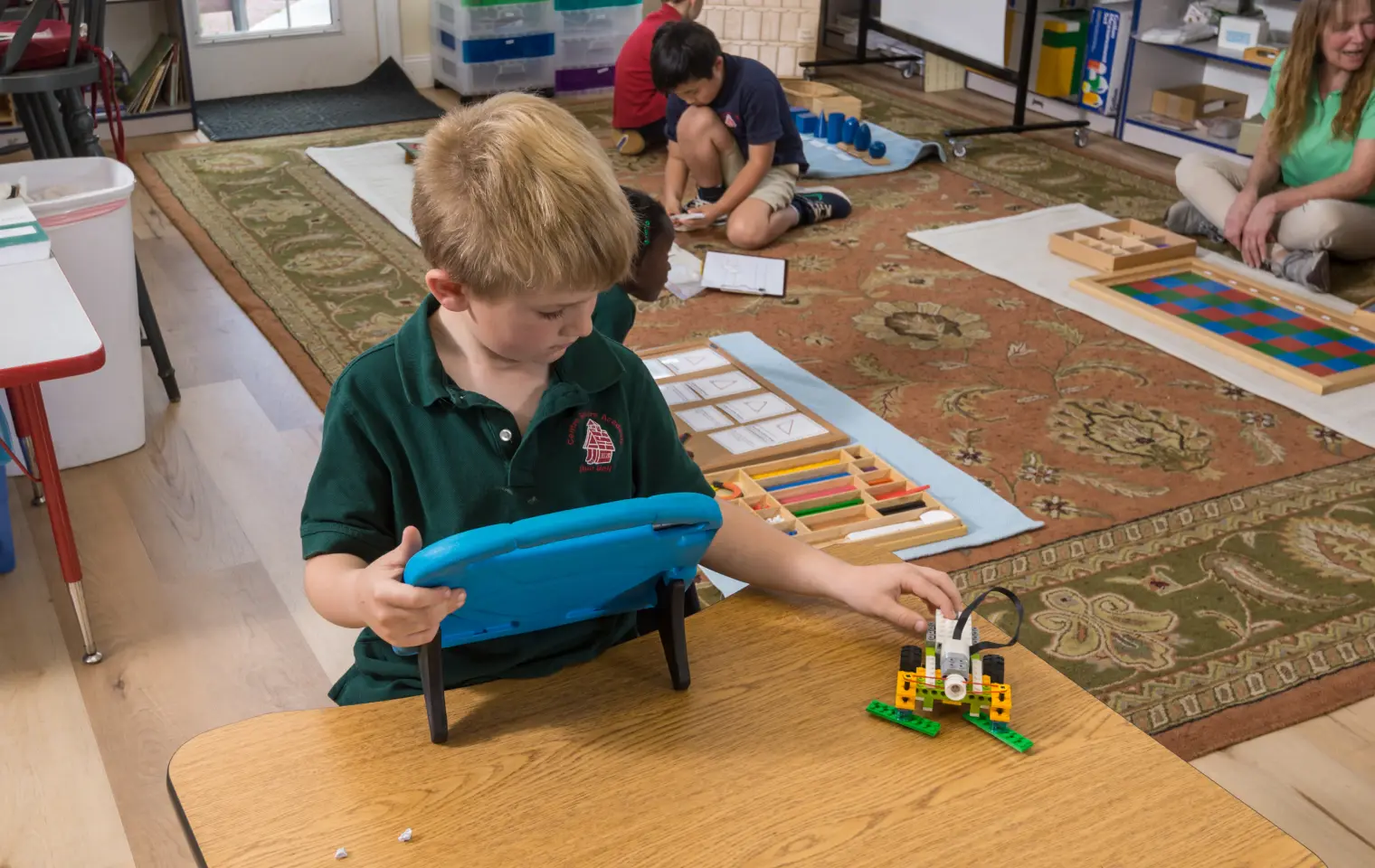 A boy in a green polo shirt holds a blue tablet case while looking at a small LEGO robot on a wooden table, with other children seated on a patterned rug in the background.