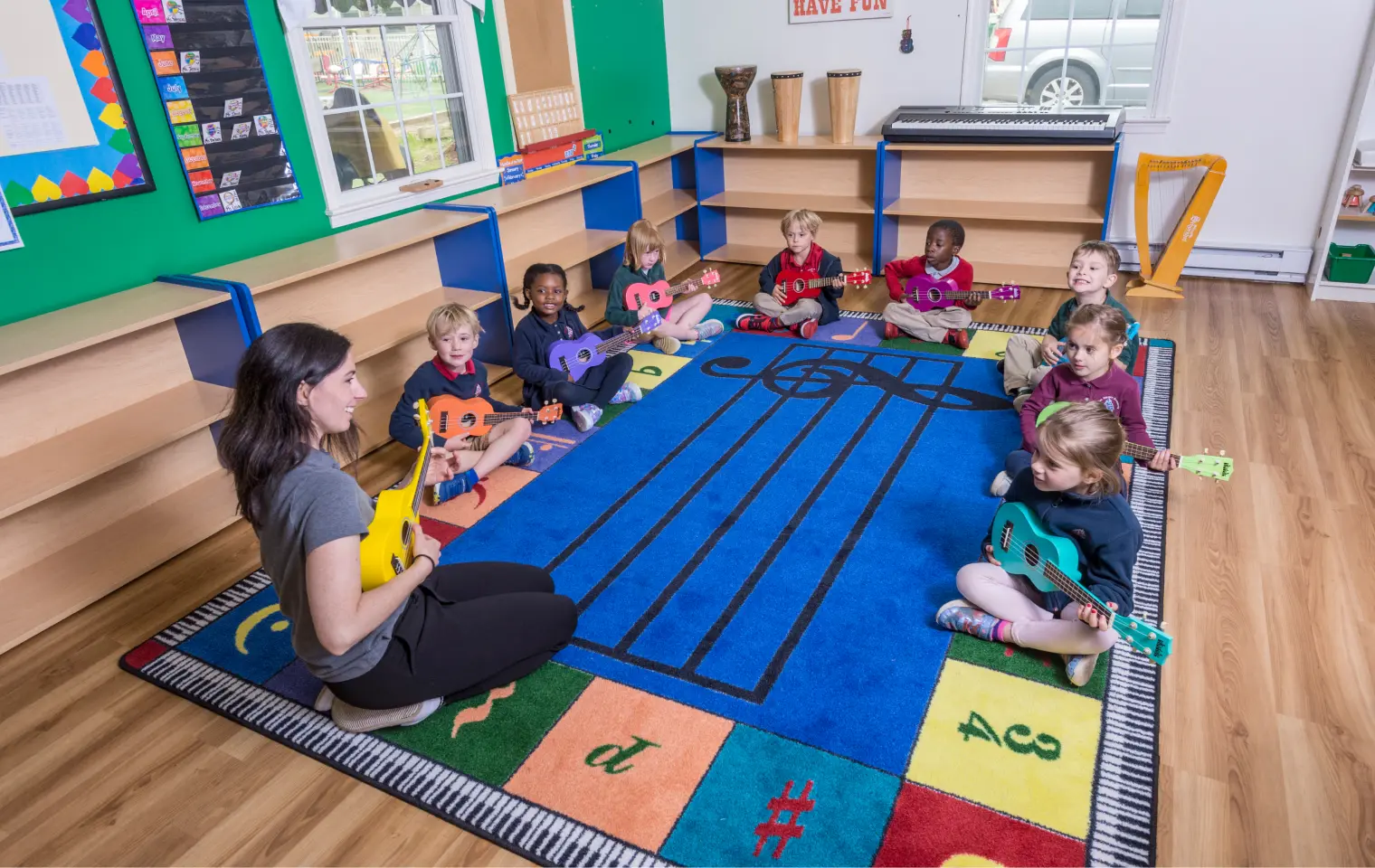 A woman sits on a music-themed rug playing a yellow ukulele while eight children seated in a circle hold small colored ukuleles.