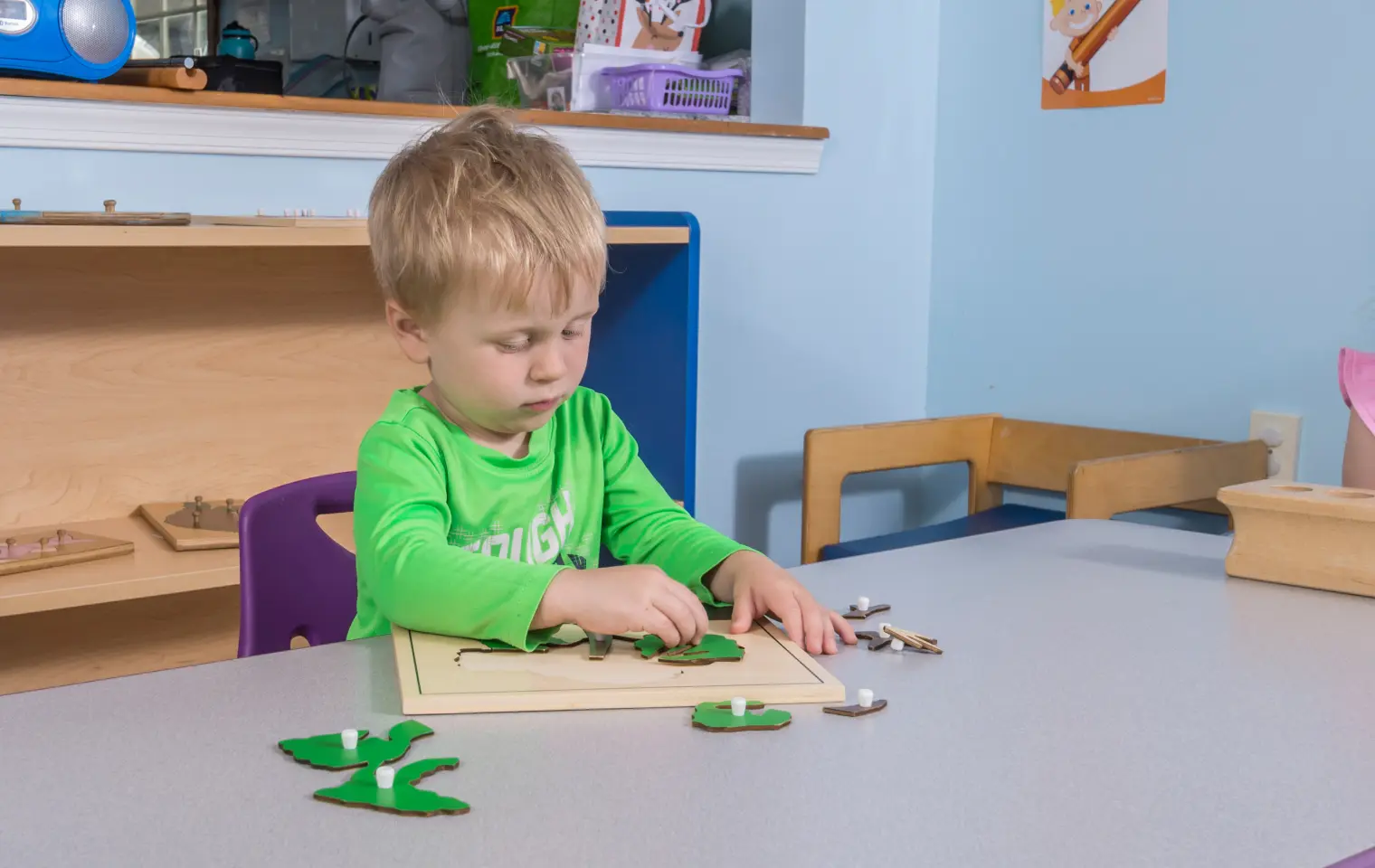 A young boy in a green shirt sits at a table and places wooden puzzle pieces onto a board.