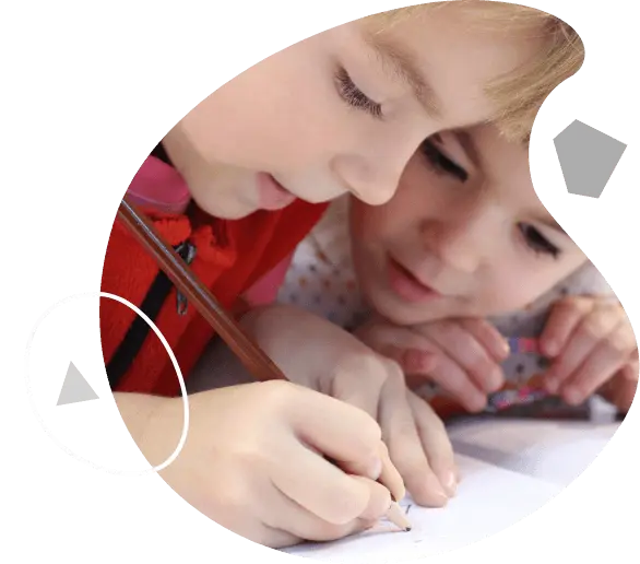 Two young children lean over a table as one holds a pencil and draws on paper while the other watches.