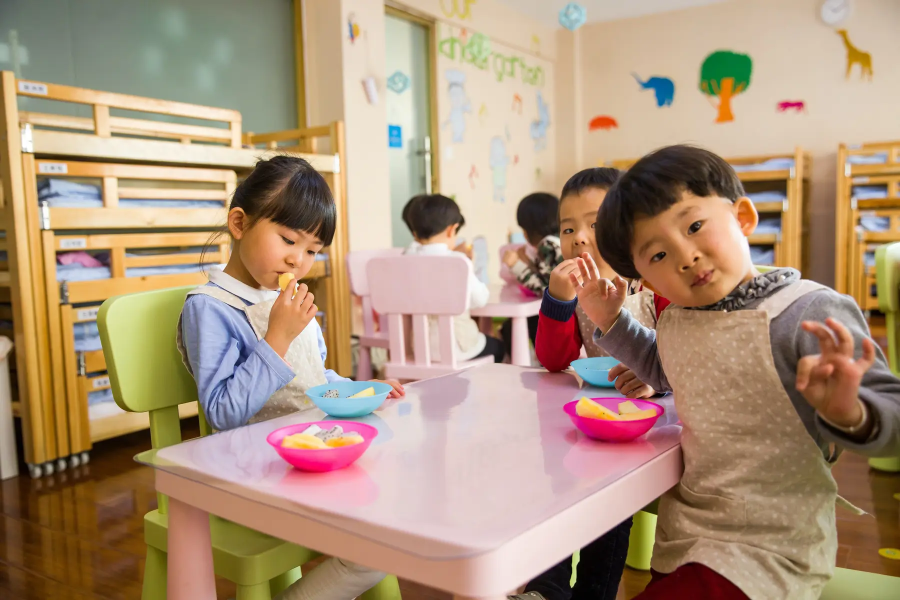 Children sit at a pink table eating fruit from colorful bowls in a room with wooden bunk beds and animal wall decorations.