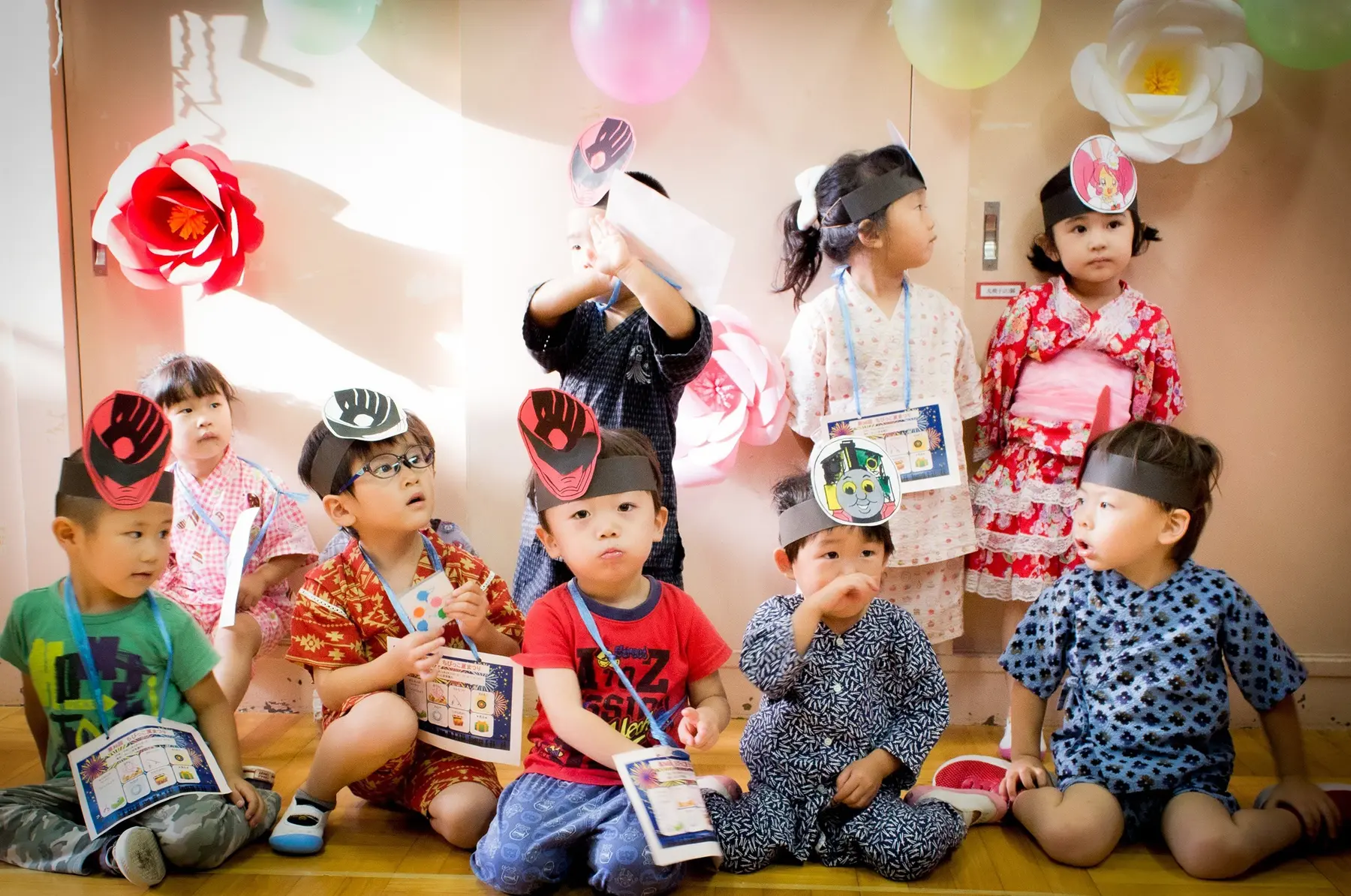 Ten children sit and stand indoors wearing paper headbands and lanyards with printed cards, some in patterned yukata, near paper flowers and balloons.