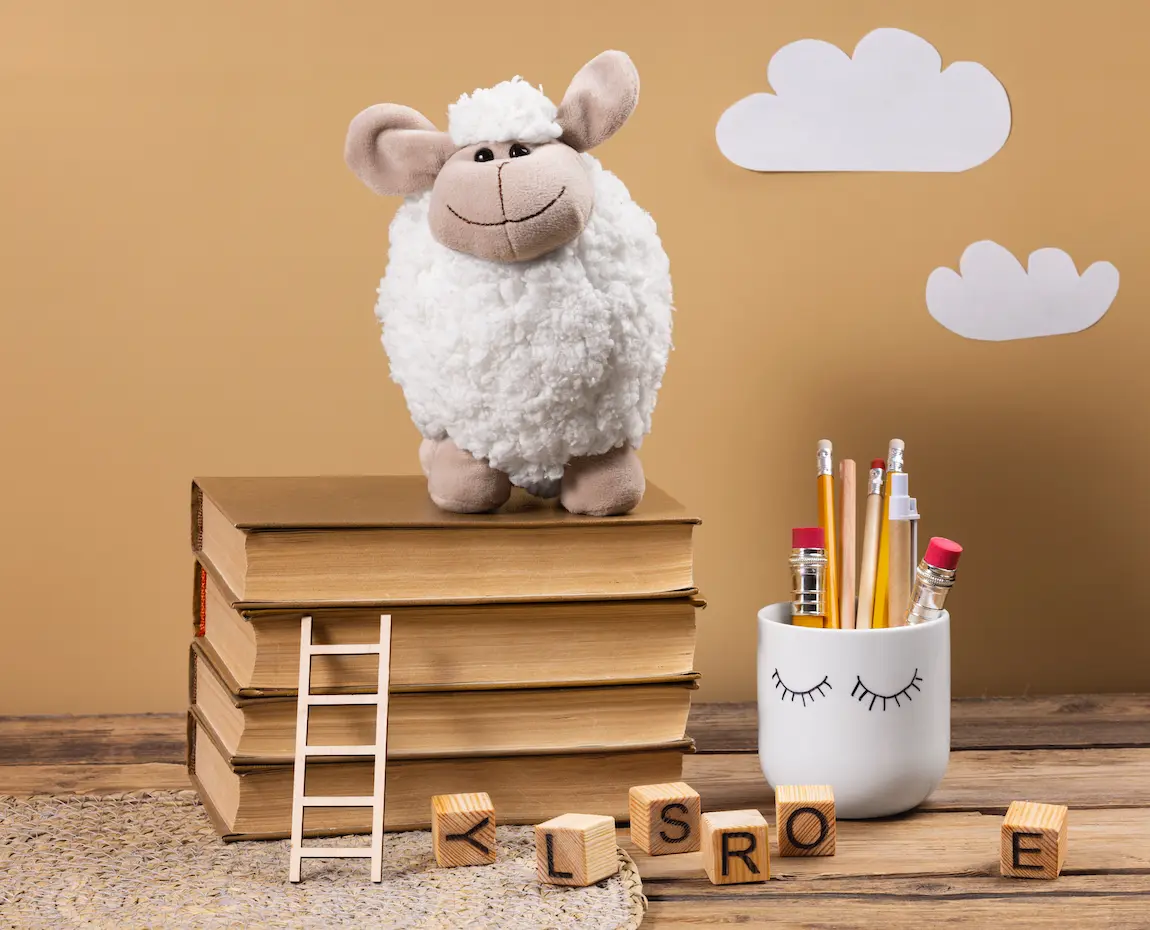 A white sheep plush toy stands on a stack of brown books next to a small wooden ladder, alphabet blocks, and a pen holder with pencils on a wooden table.