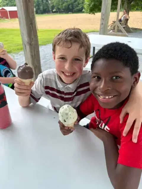 Two school-aged boys are smiling at the camera while holding ice cream cones at a white table outdoors, with another child partially visible and an adult in the background.