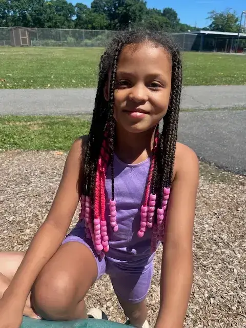 A young girl with braided hair and pink beads is smiling at the camera while outdoors on a sunny day.