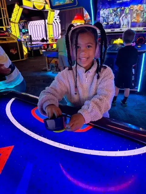A young child with braided hair and headphones smiles at the camera while playing an air hockey game in an arcade, with another child and various arcade machines in the background.
